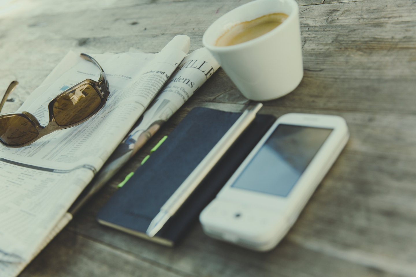 A newspaper, sunglasses, notebook and phone on a wooden table.