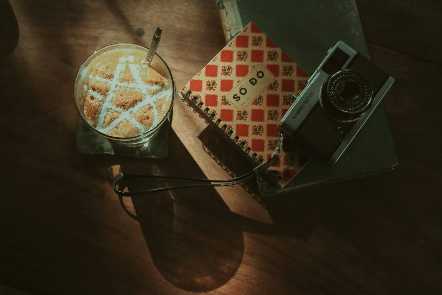 A glass of foamy coffee sits beside a notebook and camera.